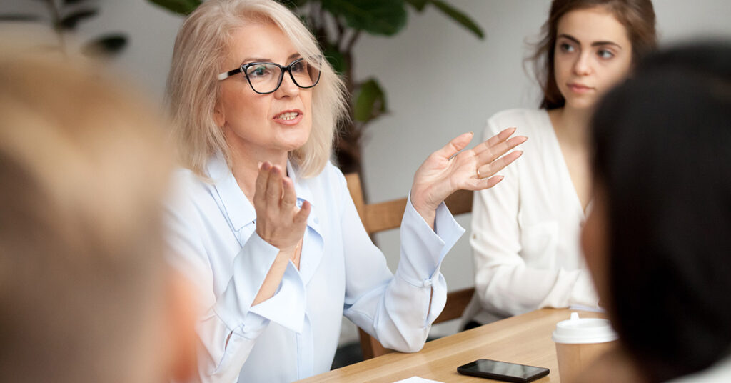 Older white woman speaking to others around a table.