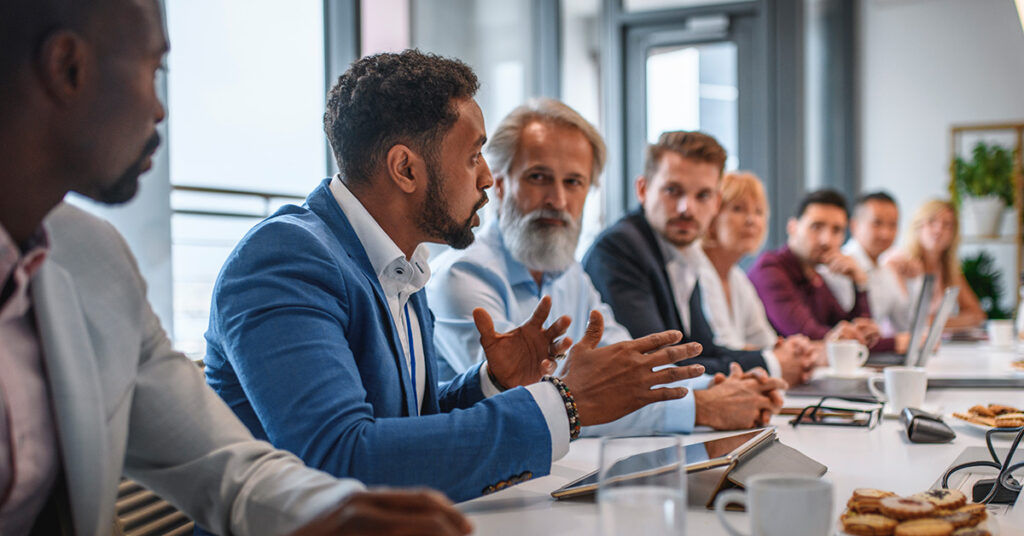 Man speaking to group around a long conference table.