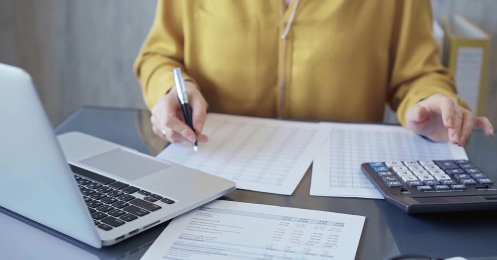 Person working with printed spreadsheets and a calculator in front of a laptop.